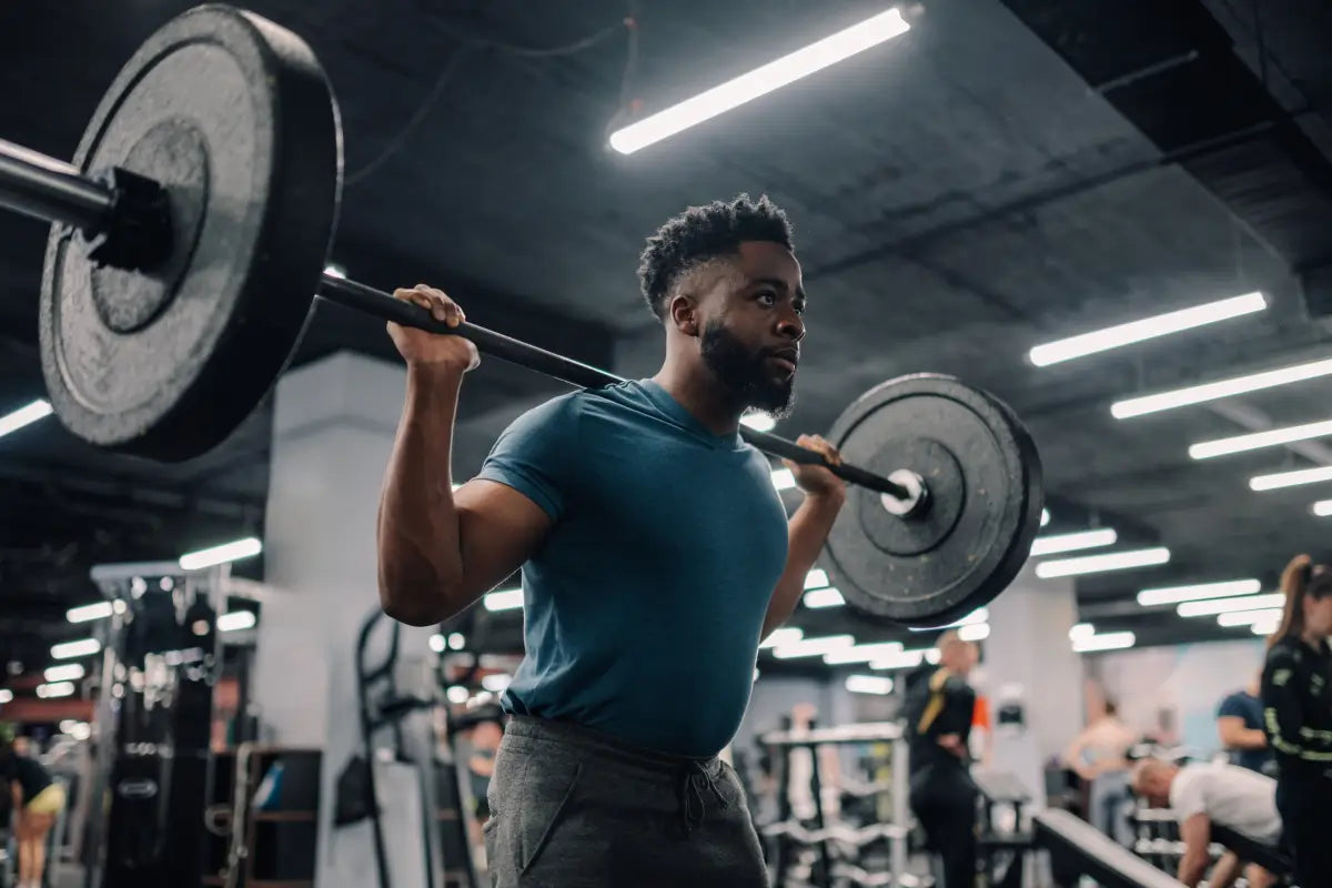 Person performing barbell shoulder press in gym with bright overhead lighting, demonstrating strength training and metabolic work